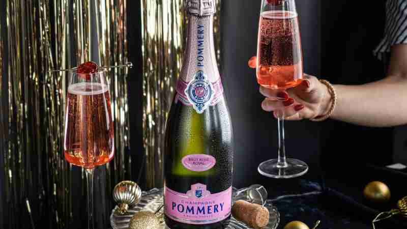 A glamorous bottle of Pommery Brut Rose sits on top a table with confetti in the background and a making merry party going on with champagne poured